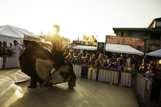 Jordan Thomas, 16, of Orange County hangs on tight while trying to beat eight seconds on a mechanical bull in front of the stand Wine Slushies at The Great New York State Fair. Onlookers smile and hold other fair refreshments as they cheer on Thomas.